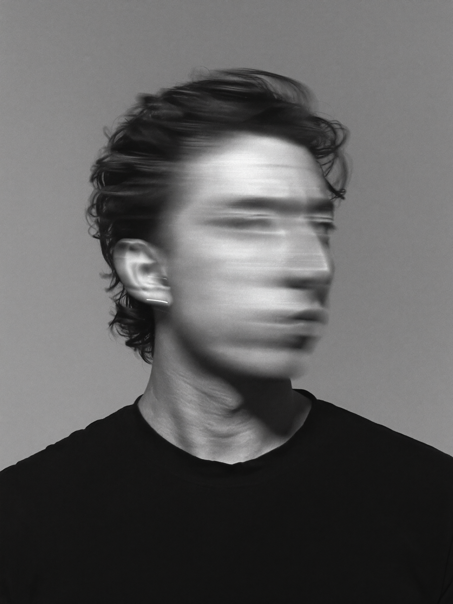 Black-and-white portrait of Efe Aydınalp against a plain grey backdrop, his face blurred by movement during a long exposure while his hair and shoulders stay sharp.